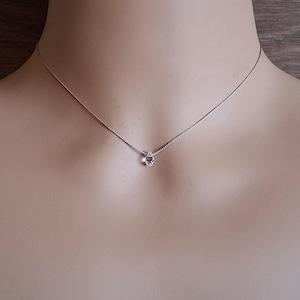 May include: A delicate silver-toned chain necklace featuring a single, small, clear, faceted gemstone pendant centered on the strand. The necklace rests against the neck, showcasing a thin, smooth metal chain and a minimalist, sparkling stone.