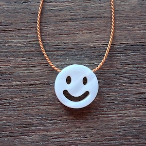 May include: A white, circular pendant necklace featuring a smiley face design. The pendant is strung on a thin, gold-colored cord. The necklace is displayed on a dark wood surface.