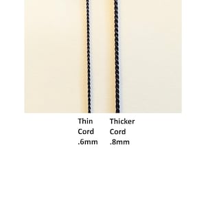 May include: Two black twisted cords displayed vertically. The left cord is labeled Thin Cord .6mm and the right cord is labeled Thicker Cord .8mm. The cords have a uniform, spiraled texture and are positioned side by side against a plain background.