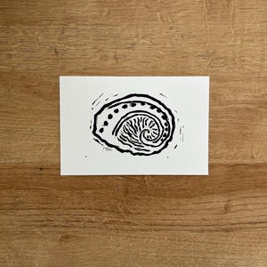 May include: A black and white linocut print of a spiral design on white paper. The print features a central spiral surrounded by a dotted line and a border of small, irregular shapes. The artwork is displayed on a wooden surface.