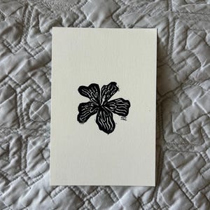 Puede incluir: Un grabado en lin&oacute;leo en blanco y negro de una flor con seis p&eacute;talos. La flor est&aacute; centrada en un trozo rectangular de papel blanquecino. La impresi&oacute;n tiene un aspecto texturizado y est&aacute; sobre un fondo acolchado gris.