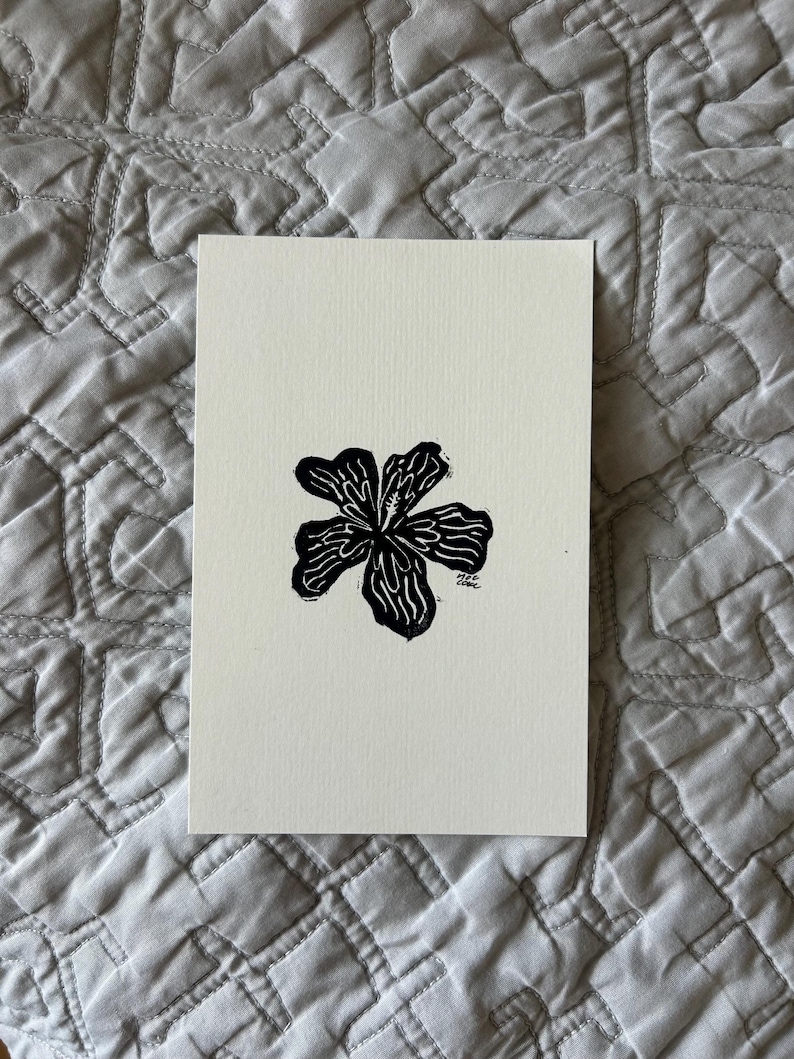 Puede incluir: Un grabado en lin&oacute;leo en blanco y negro de una flor con seis p&eacute;talos. La flor est&aacute; centrada en un trozo rectangular de papel blanquecino. La impresi&oacute;n tiene un aspecto texturizado y est&aacute; sobre un fondo acolchado gris.