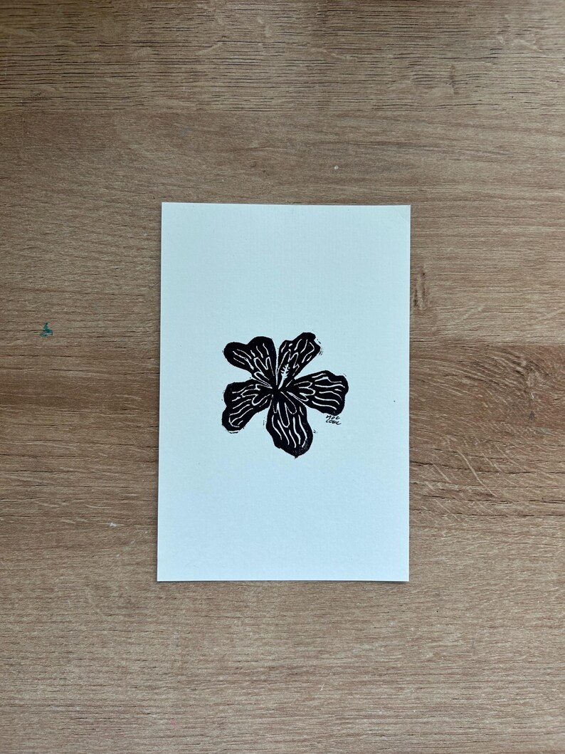 Puede incluir: Un grabado en lin&oacute;leo en blanco y negro de una flor con cinco p&eacute;talos. La flor est&aacute; centrada en un papel verde azulado claro. La impresi&oacute;n est&aacute; sobre una superficie de madera.