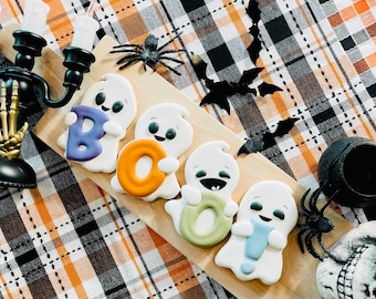 May include: Four white ghost-shaped cookies arranged on a light wood serving board. Each ghost holds a letter, spelling out the word BOO! with a purple B, orange O, green O, and light blue exclamation point. The cookies feature black eyes and mouths.