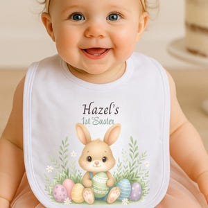 May include: A white baby bib with the text "Hazel's 1st Easter" and a cartoon bunny surrounded by Easter eggs. The bib is worn by a baby with a floral headband. A cake with flowers is in the background.