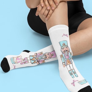 Może przedstawiać: Białe skarpetki z kolorową ilustracją postaci Dziadka do Orzechów trzymającego bęben i pudełko z prezentem. Dziadek do Orzechów ma na sobie niebieski kapelusz i różowo-złoty strój. Skarpetki mają czarne palce i pięty.