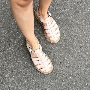 May include: A pair of light pink leather sandals with a caged design and buckle closures. The sandals are worn on a person's feet, standing on a gray asphalt surface.