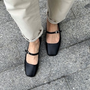 May include: A pair of black leather mary jane shoes with a buckle strap. The shoes are worn by a person wearing beige pants.