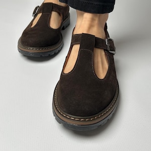 Puede incluir: Primer plano de zapatos marrones de ante con correa en T y cierre de hebilla. Los zapatos tienen punta redonda y suela gruesa y texturizada. El fondo es una superficie blanca lisa.