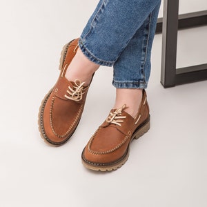 Op de afbeelding: Een paar bruine leren bootschoenen met witte veters. De schoenen worden gedragen door iemand die een blauwe jeans draagt.