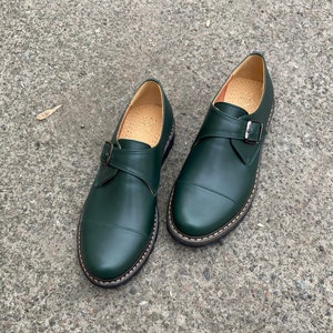 May include: A pair of dark green leather shoes with a single buckle strap. The shoes have a rounded toe and a stitched edge.