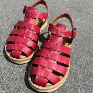 Puede incluir: Un par de sandalias de cuero rojo con un dise&ntilde;o tejido. Las sandalias tienen un cierre de hebilla y una suela beige. Las sandalias miden aproximadamente 20 cm de largo.