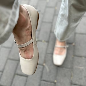 May include: A pair of off-white leather shoes with a strap across the top. The shoes have a square toe and a low heel.
