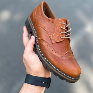Pu&ograve; includere: Scarpa Oxford in pelle marrone con perforazioni decorative e suola spessa nera. La scarpa ha lacci colorati chiari e una tomaia in pelle strutturata. La scarpa &egrave; tenuta in mano.