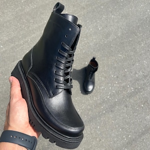 Puede incluir: Botas de combate de cuero negro con diseño de cordones y suela gruesa. Las botas tienen punta redondeada y una lengüeta en la parte trasera. Una segunda bota es visible en el fondo. Las botas miden aproximadamente 18 cm de alto.