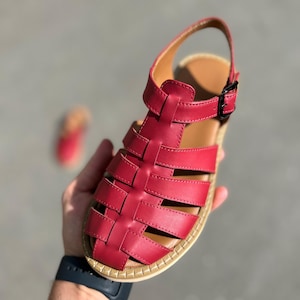 Puede incluir: Sandalia de cuero rojo con dise&ntilde;o tejido. La sandalia tiene cierre de hebilla y plantilla marr&oacute;n. La suela tiene un borde beige. La sandalia es sostenida por una mano.