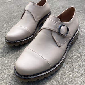 K&ouml;nnte beinhalten: Ein Paar beige Lederschuhe mit Schnallenverschluss. Die Schuhe haben eine runde Spitze und eine dicke, strukturierte Sohle. Die Schuhe sind etwa 25 cm lang.