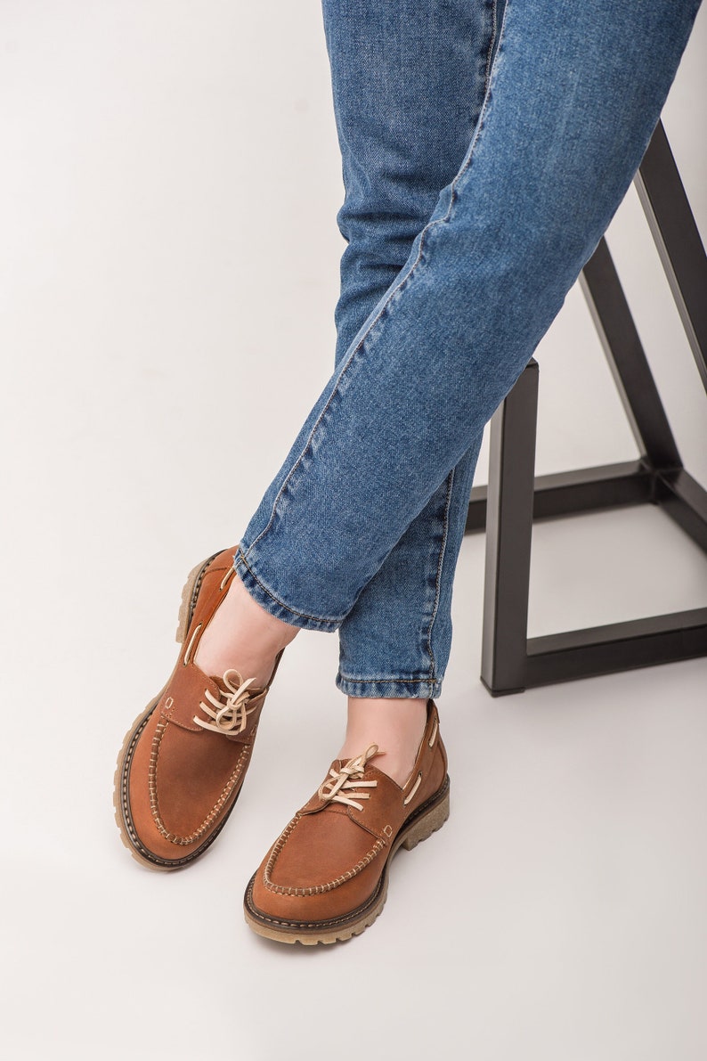 Op de afbeelding: Een paar bruine leren bootschoenen met witte veters. De schoenen worden gedragen door iemand die een blauwe jeans draagt.