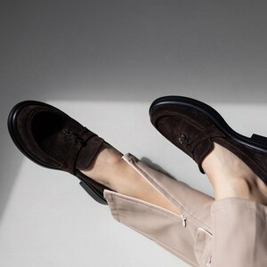 May include: A pair of brown suede loafers with a zipper detail on the side. The loafers are worn by a person wearing beige pants.