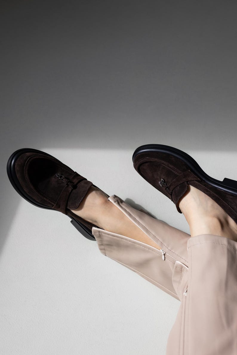 May include: A pair of brown suede loafers with a zipper detail on the side. The loafers are worn by a person wearing beige pants.