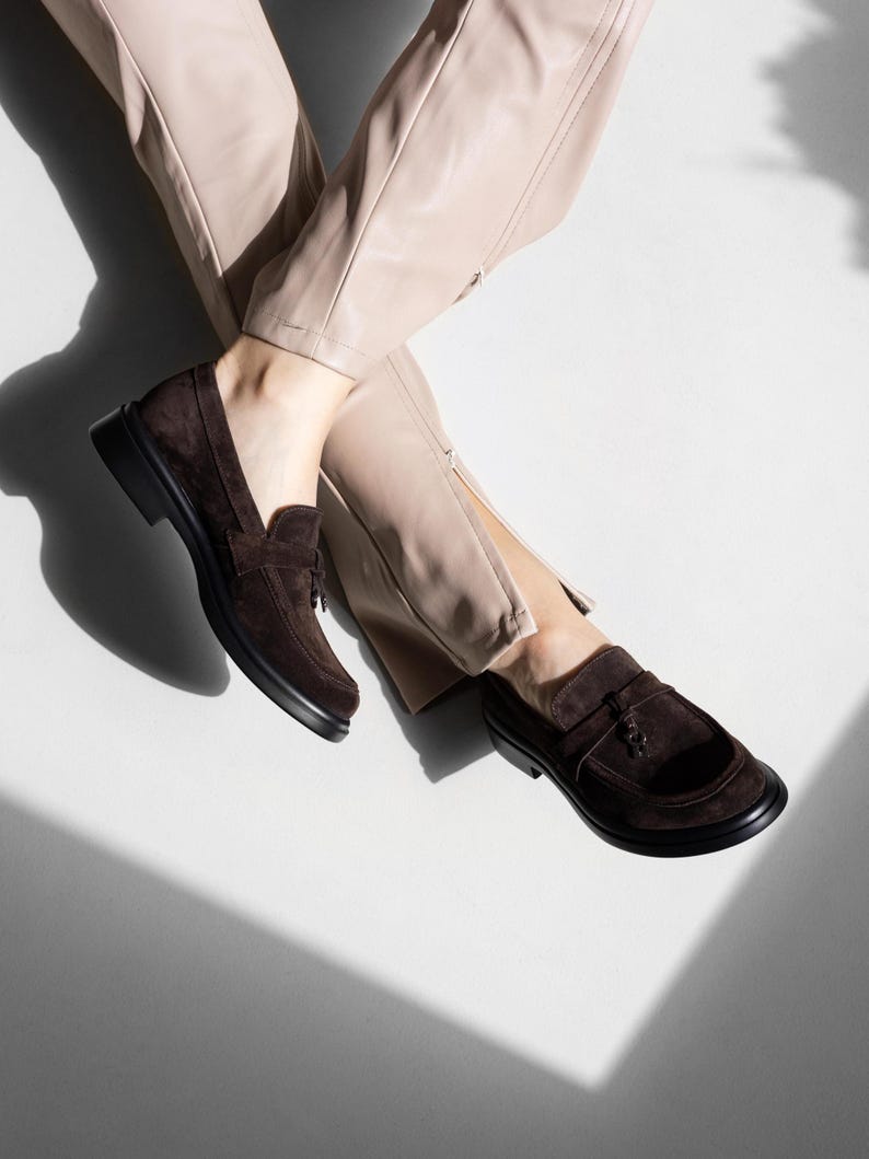 May include: A pair of brown suede loafers with tassels. The loafers are worn by a person wearing beige pants.