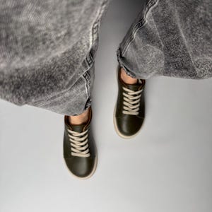 May include: A pair of dark green leather low-top sneakers with white laces. The sneakers are worn by a person wearing gray jeans.