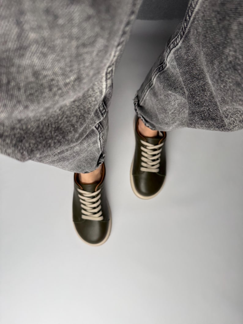 May include: A pair of dark green leather low-top sneakers with white laces. The sneakers are worn by a person wearing gray jeans.
