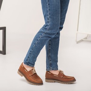 Op de afbeelding: Bruine leren bootschoenen met witte veters. De schoenen hebben een bruine rubberen zool en worden gedragen met een blauwe jeans.
