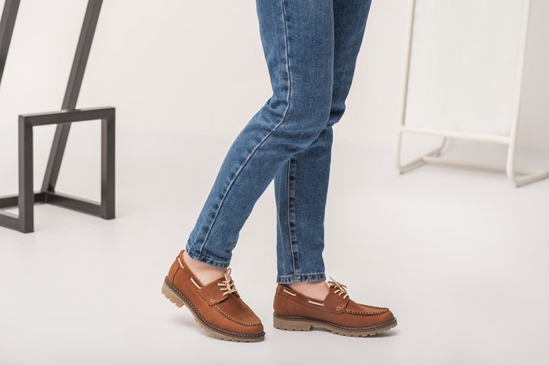 Op de afbeelding: Bruine leren bootschoenen met witte veters. De schoenen hebben een bruine rubberen zool en worden gedragen met een blauwe jeans.