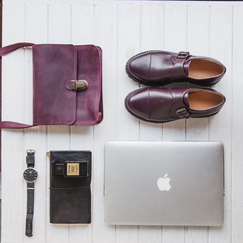 K&ouml;nnte beinhalten: Eine flache Anordnung aus Laptop, Ledertasche, Uhr, einem kleinen schwarzen Notizbuch und einem Parf&uuml;mflakon. Der Laptop ist silberfarben mit einem wei&szlig;en Apple-Logo. Die Tasche ist burgunderfarben mit einer Messing-Schnalle. Die Uhr ist silberfarben mit einem schwarzen Band. Das Notizbuch ist schwarz mit einem braunen Lederbezug. Der Parf&uuml;mflakon ist aus klarem Glas mit einem goldenen Verschluss.
