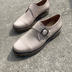 K&ouml;nnte beinhalten: Ein Paar beige Lederschuhe mit Schnallenverschluss und runder Spitze. Die Schuhe haben eine dicke, strukturierte Sohle und eine dekorative Naht am Rand. Die Schuhe stehen auf einer Betonoberfl&auml;che.