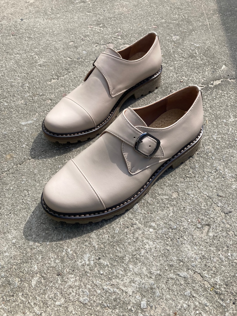 K&ouml;nnte beinhalten: Ein Paar beige Lederschuhe mit Schnallenverschluss und runder Spitze. Die Schuhe haben eine dicke, strukturierte Sohle und eine dekorative Naht am Rand. Die Schuhe stehen auf einer Betonoberfl&auml;che.