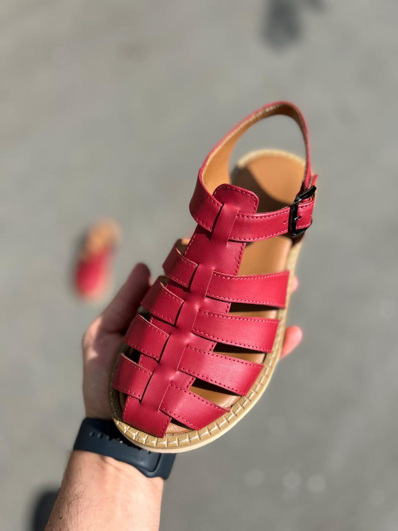 Puede incluir: Sandalia de cuero rojo con dise&ntilde;o tejido. La sandalia tiene cierre de hebilla y plantilla marr&oacute;n. La suela tiene un borde beige. La sandalia es sostenida por una mano.