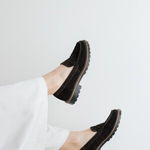 May include: A pair of black suede loafers with a chunky sole. The loafers are worn by a person with white pants.
