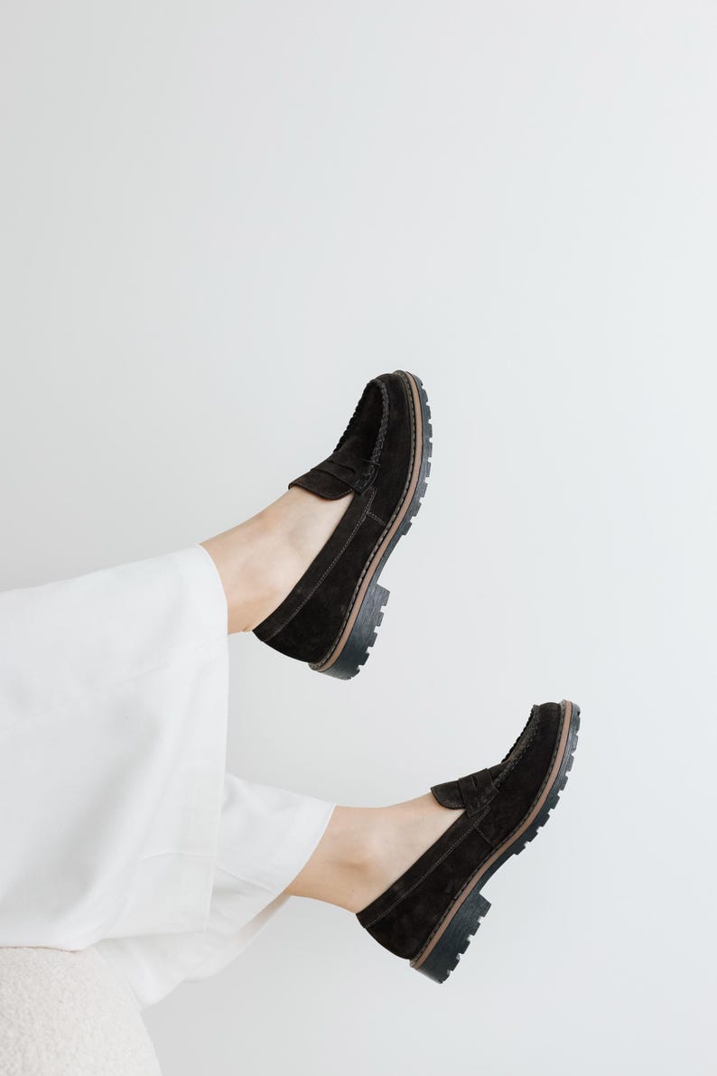 May include: A pair of black suede loafers with a chunky sole. The loafers are worn by a person with white pants.