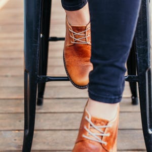May include: A pair of brown leather lace-up shoes with white laces. The shoes are worn by a person sitting on a black metal stool.