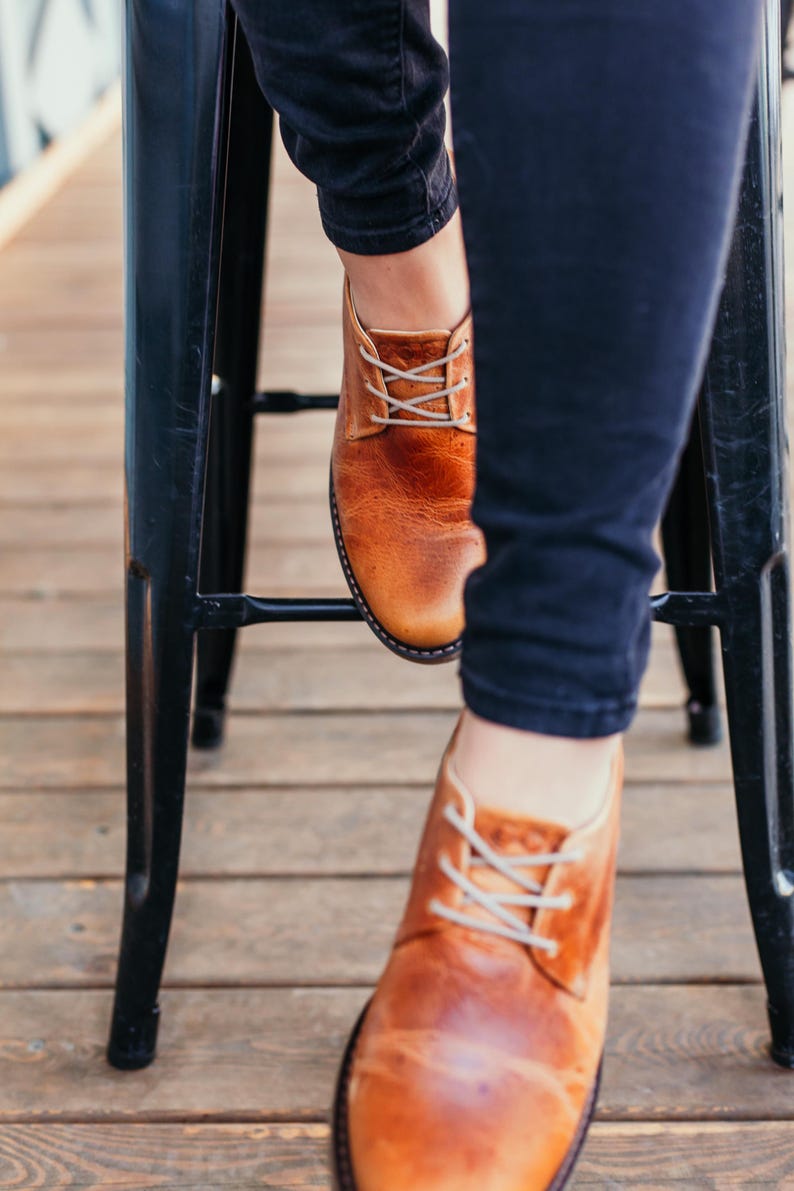 May include: A pair of brown leather lace-up shoes with white laces. The shoes are worn by a person sitting on a black metal stool.