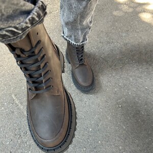 May include: Brown leather lace-up boots with a thick black rubber sole. The boots are worn with denim jeans.