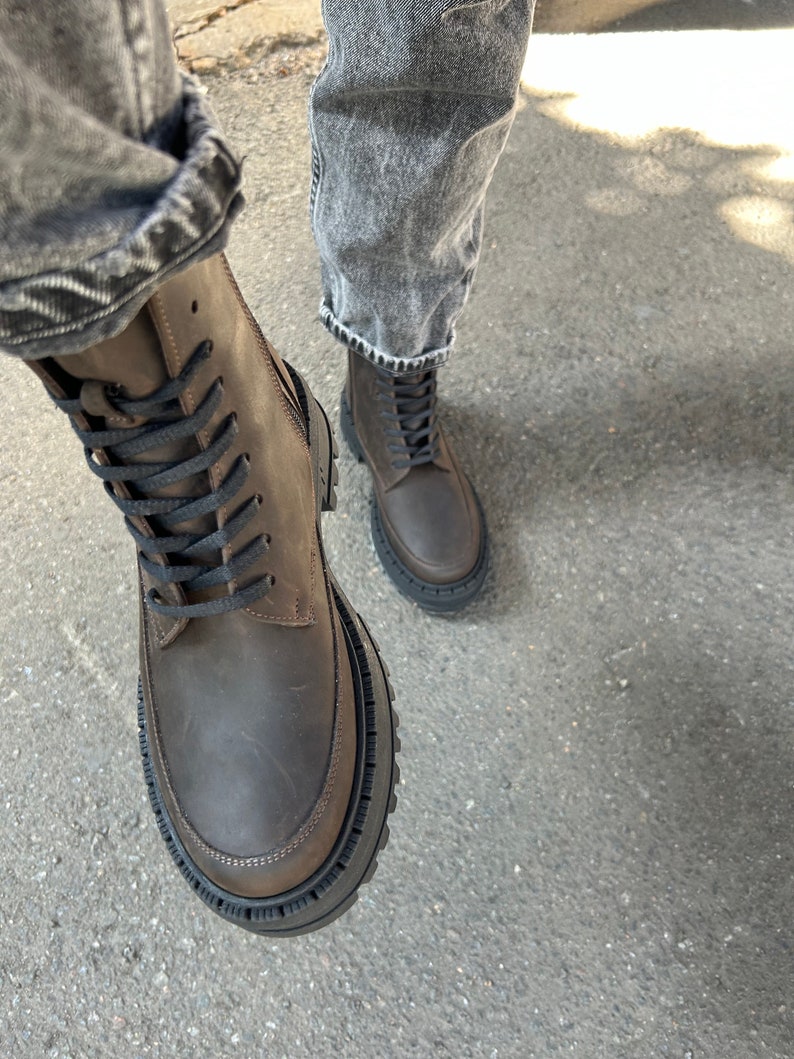 May include: Brown leather lace-up boots with a thick black rubber sole. The boots are worn with denim jeans.