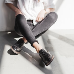May include: A person wearing a white button-down shirt and black jeans sits with their legs crossed. They are wearing black leather lace-up shoes.