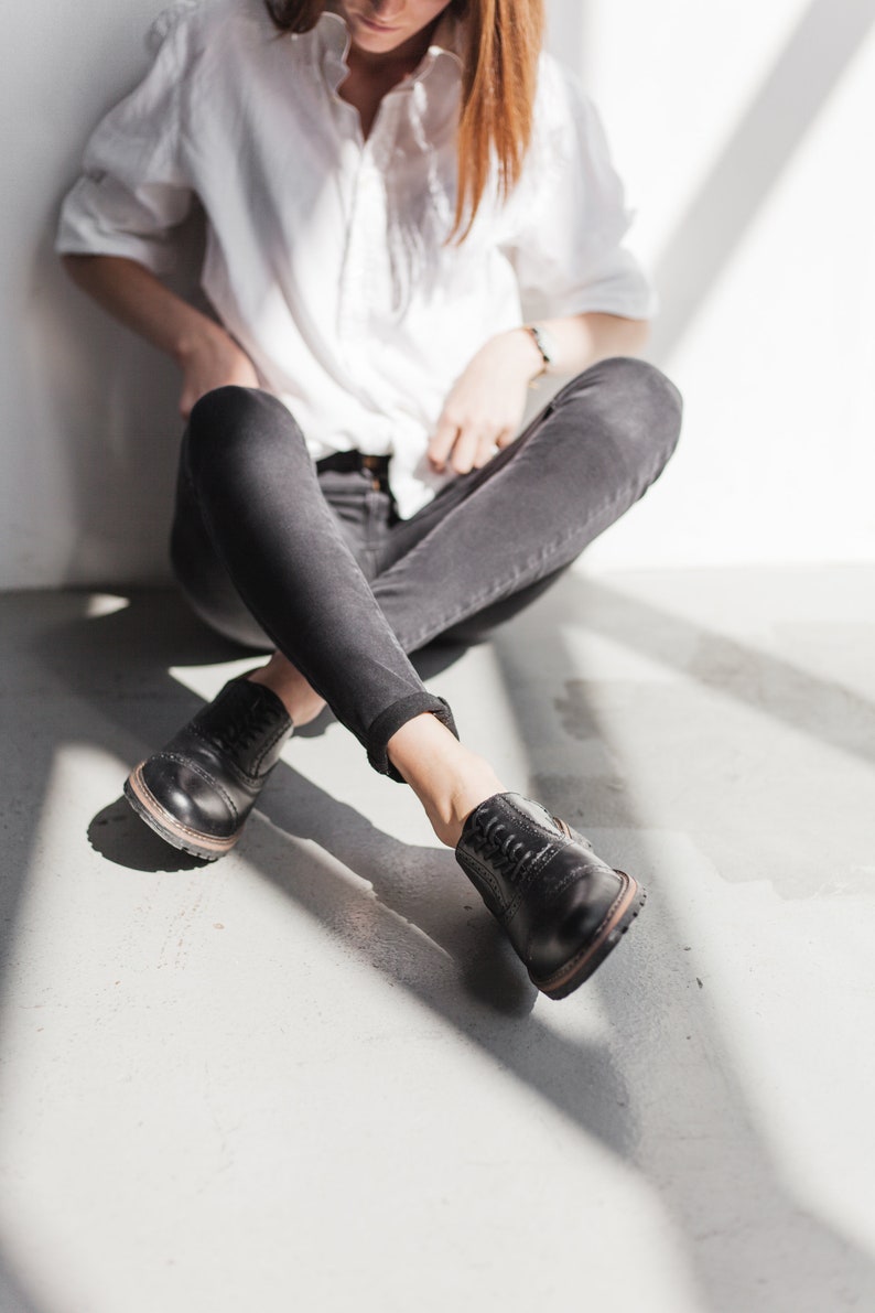 May include: A person wearing a white button-down shirt and black jeans sits with their legs crossed. They are wearing black leather lace-up shoes.