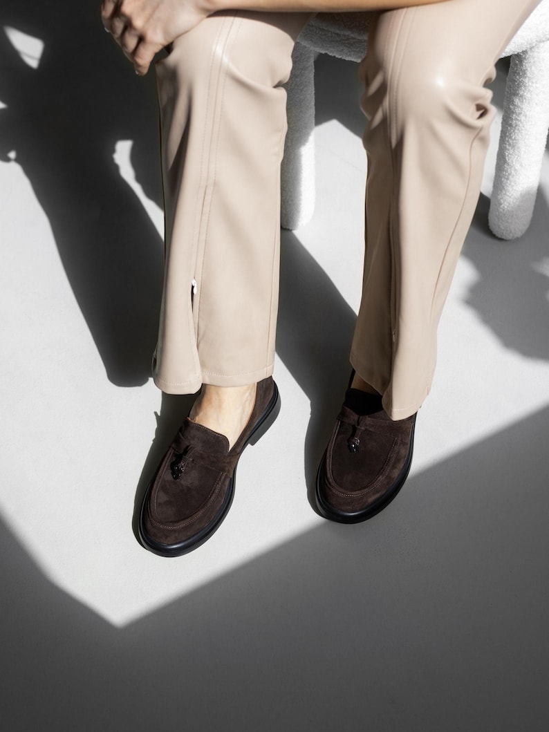 May include: A pair of brown suede loafers with tassel accents. The loafers have a dark brown sole and are worn with beige pants.