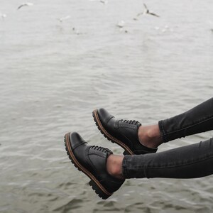 May include: A pair of black leather oxford shoes with laces. The shoes are worn by a person sitting with their legs dangling over the edge of a pier. The water is visible in the background.