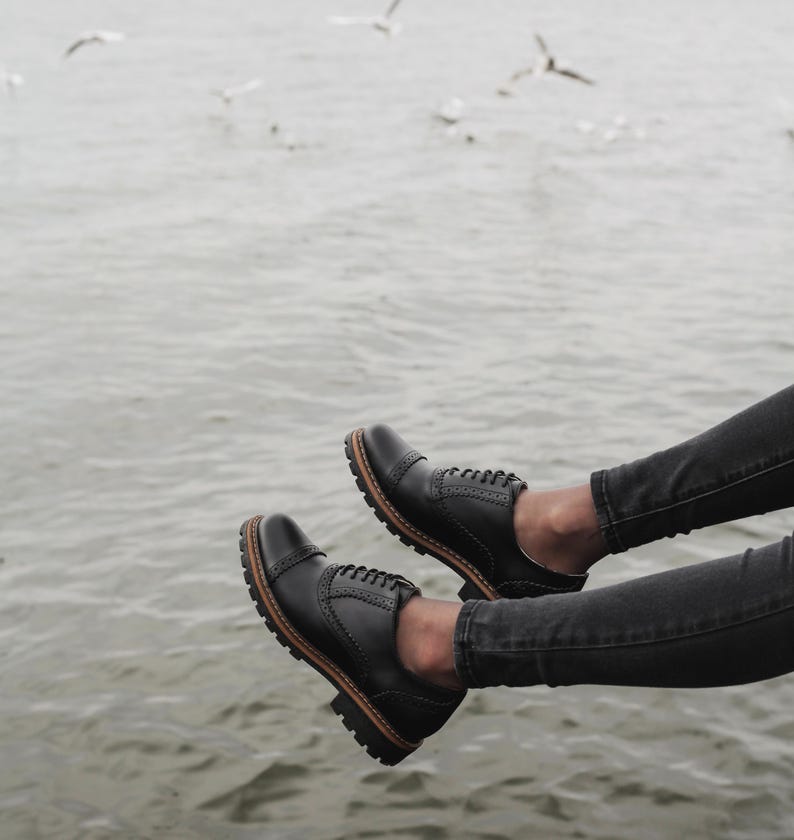 May include: A pair of black leather oxford shoes with laces. The shoes are worn by a person sitting with their legs dangling over the edge of a pier. The water is visible in the background.