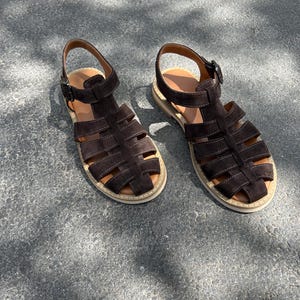 Pu&ograve; includere: Un paio di sandali in pelle scamosciata marrone scuro con un design a gabbia. I sandali hanno un cinturino alla caviglia con fibbia e una suola colorata chiara. Le scarpe sono esposte su una superficie grigia, mostrando il loro design.