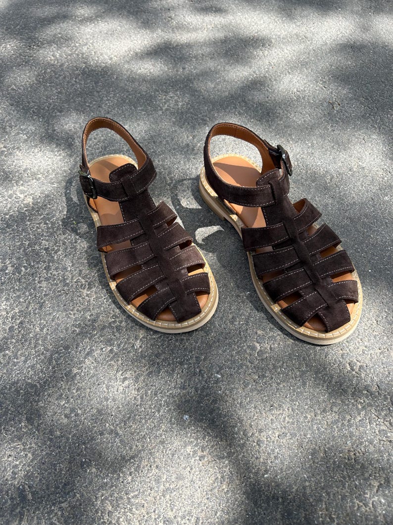 Pu&ograve; includere: Un paio di sandali in pelle scamosciata marrone scuro con un design a gabbia. I sandali hanno un cinturino alla caviglia con fibbia e una suola colorata chiara. Le scarpe sono esposte su una superficie grigia, mostrando il loro design.