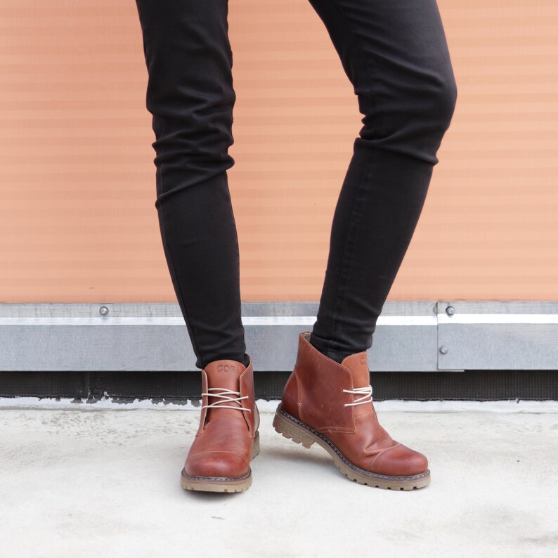 May include: Brown leather ankle boots with laces. The boots have a chunky sole and are worn with black pants.