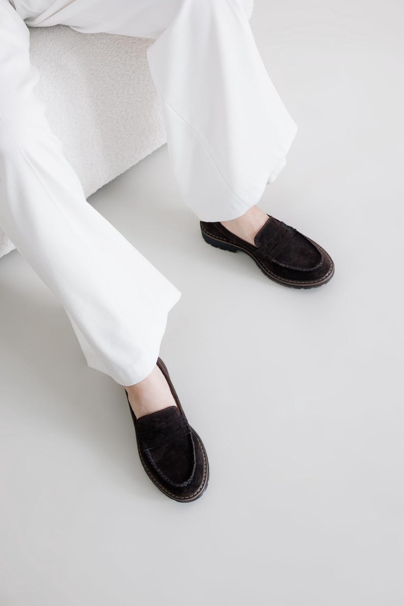 May include: A pair of brown suede loafers with a tassel detail. The loafers are worn by a person wearing white pants.