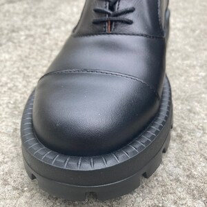 May include: Close-up of the black leather toe of a lace-up shoe with a thick, textured rubber sole.