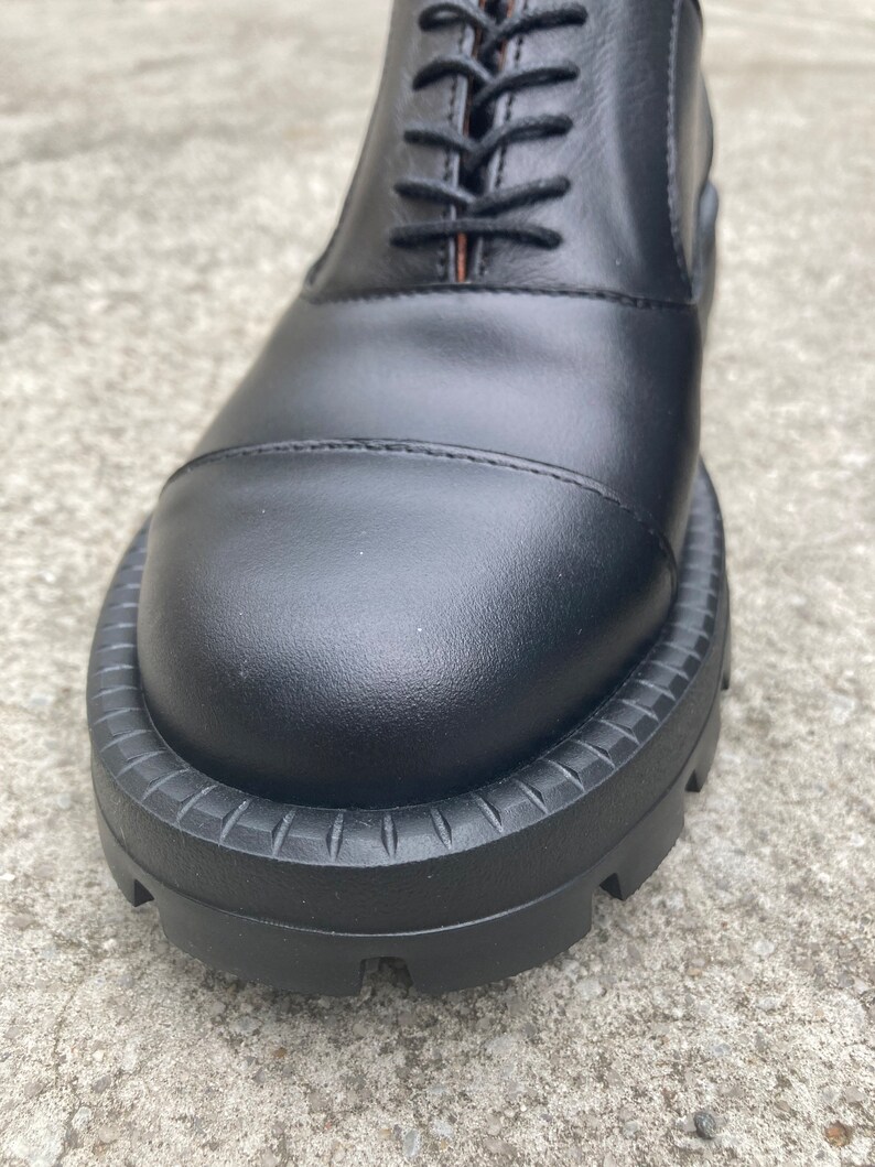 May include: Close-up of the black leather toe of a lace-up shoe with a thick, textured rubber sole.